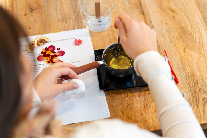 Woman melting natural soy wax in a saucepan, adding essential oils, and incorporating dried flowers to make scented candles.