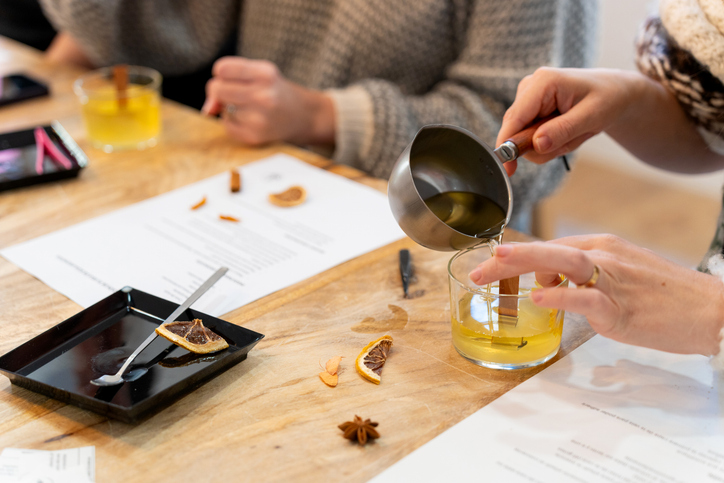 A woman's hands pouring essential oil-infused soy wax into a glass jar with a wooden wick during a candle-making workshop